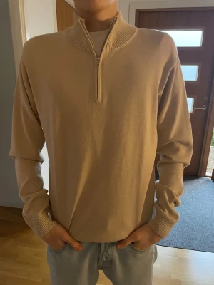 Beige stickad tröja med half zip - Snygg beige stickad tröja med half zip och ribbad krage. Tröjan har långärmade ärmar och en stilren design som passar perfekt till jeans. Materialet är mjukt och bekvämt, och den neutrala färgen gör den enkel att matcha med olika outfits.
