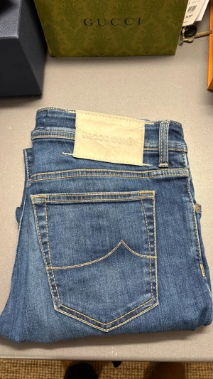 Blå jeans från Jacob Cohen - Snygga blå jeans från Jacob Cohen med klassisk femficksdesign och kontrastsömmar. Jeansen har en ljus läderpatch bak i midjan och insidan är fodrad med ett randigt tyg i blått, rött och vitt. Passformen är rak och materialet är mjukt denim.