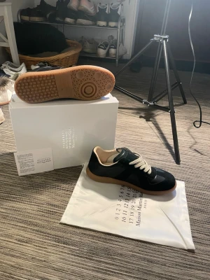 Maison Margiela svarta sneakers - Säljer ett par snygga maison margiela Gats! Skorna är i mycket bra skick, använda några få gånger. Inga defekter! Enda defekten är att boxen är lite buktig. Storlek 43 men passar 42. Allting på bilderna tillkommer dvs box, dustbag, papper och kort. Om ni har några frågor så är de bara att höra av er😊