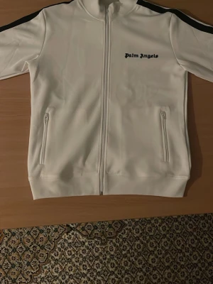 Palm Angels - Säljer en clean vit track jacket från Palm Angels med svarta ränder längs axlarna och logga på bröstet. Jackan har hel dragkedja framtill, två fickor med dragkedja och hög krage. Perfekt för en sportig och stilren look.