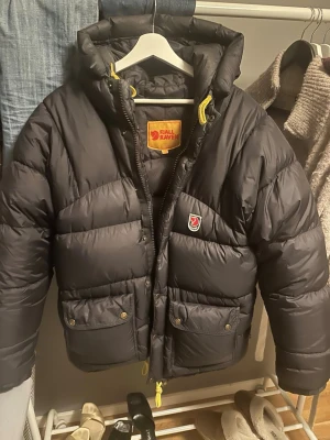 Svart dunjacka från Fjällräven XXS - Säljer en svart dunjacka från Fjällräven i storlek XXS. Jackan har huva, gula snören och två stora fickor med knappar framtill. Perfekt för kalla dagar och har en klassisk Fjällräven-logga på bröstet. Stängs med dragkedja och tryckknappar.