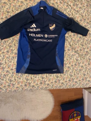 Blå IFK Norrköping träningströja Adidas - Säljer en blå träningströja från IFK Norrköping med tryck på bröstet och ryggen, nummer 2 i nacken och flera sponsorer. Tröjan har halv dragkedja, lång ärm och är gjord i polyester. Perfekt för dig som gillar fotboll och vill ha något snyggt från Peking.