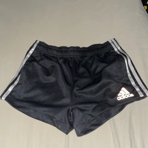 Svarta Adidas Climalite shorts - Säljer ett par svarta Adidas shorts med silvriga ränder längs sidorna och klassisk Adidas-logga framtill. Modellen är Climalite, vilket gör dem extra sköna och luftiga för träning. Elastisk midja och lätt material i polyester.