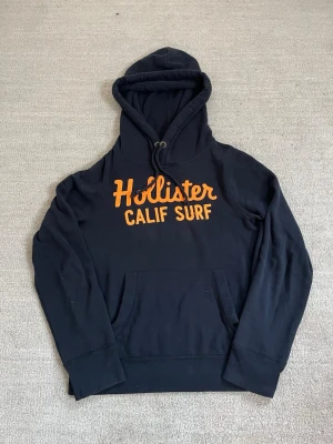 Hollister Hoodie - Tja! Säljer nu denna feta hoodie från Hollister! Den är i riktigt bra skick och har inga defekter. Den har ett snyggt tryck på tröjan som ger den en lite ”Archive” stil. Storleken är S men passar även XS. Skriv ifall du funderar på något eller har en fråga, och pris kan diskuteras!