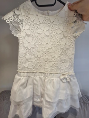 Vit blommig barnklänning med spets - Superfin vit barnklänning med blommigt spetsmönster upptill och volangkjol i lager. Klänningen har korta ärmar med spetsdetaljer och pärlor vid midjan. Dragkedja i ryggen och dekorativ blomma på kjolen. Perfekt för sommarens alla tillfällen!