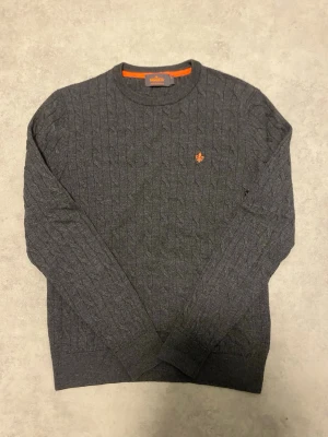 Mörkgrå stickad tröja från Morris - Snygg mörkgrå stickad tröja från Morris med kabelstickat mönster och rund hals. Tröjan är 100% merinoull. Tröjan har ett orange broderat emblem på bröstet och ribbade muddar vid ärmslut och nederkant. Perfekt för dig som gillar klassisk stil med en modern touch.