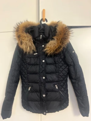 Svart pufferjacka Rockandblue med päls - Snygg svart pufferjacka från Rockandblue med fluffig päls runt huvan. Jackan har quiltade detaljer, flera dragkedjor och knappar framtill samt en patch på ärmen. Perfekt för kalla dagar med sin varma och stilrena look.lite fel på armen kolla bilder 