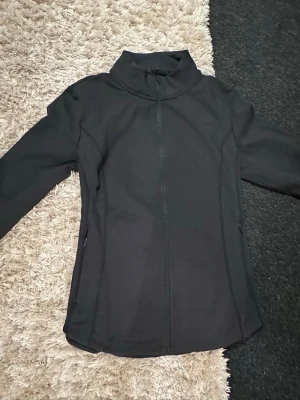 Svart träningsjacka från SOC - Svart träningsjacka från SOC i storlek L men sitter som en S tycker jag då jag brukar ha det i kläder . Jackan har hög krage, hel dragkedja framtill och är tillverkad i ett lätt och stretchigt syntetmaterial. Perfekt för träning eller utomhusaktiviteter. Enkel och stilren design utan synliga loggor på utsidan. 
