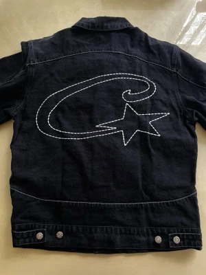 Svart jeansjacka från Corteiz - Stilren svart jeansjacka i en klassisk modell som passar till allt. Robust denim, snygga silverfärgade knappar och två bröstfickor med lock samt sidofickor. En tidlös jacka som fungerar både vår, sommar och höst.