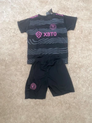 Svart och rosa Messi fotbollströja - Säljer en svart och rosa fotbollströja med tillhörande shorts från Inter Miami. Tröjan har zebramönster, klubbmärke och sponsortryck framtill samt Messi och nummer 10 på ryggen. Materialet är lätt polyester, perfekt för fotbollsträning.