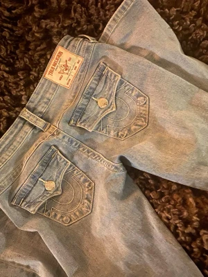 True Religion Joey Low Rise Flare Jeans - Säljer ett par ljusblå True Religion Joey jeans med låg midja och flare-ben. Klassiska jeansfickor med lock och dekorativa sömmar samt metallknappar med logga. Materialet är jeans och passformen är loose med bootcut-stil. Perfekt för dig som gillar retrovibe och detaljer.
