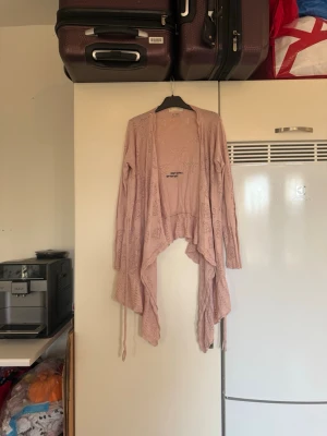 Ljusrosa stickad kofta med mönster - Säljer en ljusrosa stickad kofta med lång ärm och snyggt hålmönster i geometriska former. Koftan har omlott-design med knytband och faller snyggt framtill. Perfekt för lager-på-lager och chill stil. Superskön och mjuk känsla. Storlek 1 som enligt deras storleksguide motsvarar en S.  Mycket fint skick, jag hittar inga synliga anmärkningar eller defekter. 
