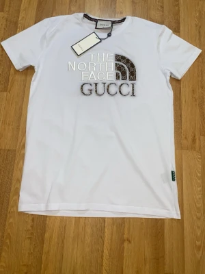 Vit t-shirt XL - Exklusiv vit t-shirt  i storlek XL. Snyggt broderad logga framtill med silvriga bokstäver och mönstrad halvcirkel med print. Klassisk rund hals och korta ärmar. Mjuk bomullskvalitet och diskret grön detalj i nederkant.