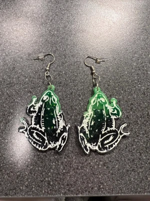 Grön svarta grod örhängen - Unik örhängen i grönt och svart. Örhängena har en cool look med vita detaljer som ska föreställa en groda, som ger en edgy vibe. Perfekt för dig som vill sticka ut med ett djurinspirerat plagg och gillar färgstarka accessoarer.