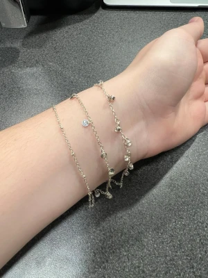 Silverfärgad armband från Claire's - Säljer ett silverfärgad armband från Claire's med enkel design och smala axelband. Armbandet har detaljer som små kristaller på ena kedjan och små brickor på andra kedjan. Perfekt för dig som gillar stilrena och diskreta plagg med en touch av glans.