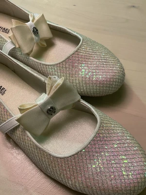 Glittriga ballerinaskor Michael Kors - Säljer ett par ballerinaskor från Michael Kors i skimrande ljus färg med nätmönster och glittereffekt. Skorna har rund tå, elastiskt band över foten och en stor cremefärgad rosett med metallogo framtill. Perfekta för dig som vill ha något extra till outfiten.