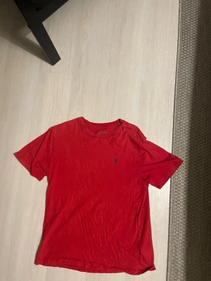 Röd t-shirt från Polo Ralph Lauren - Säljer en röd t-shirt från Polo Ralph Lauren med klassisk rund hals och korta ärmar. T-shirten har den ikoniska broderade loggan i mörkblått på bröstet. Materialet är mjuk bomull som känns skön mot huden. Perfekt för dig som gillar stilrena och tidlösa plagg. Passar om du är mellan 160-170