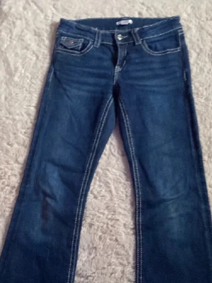 Blå bootcut jeans från Gina Tricot - Snygga blå jeans från Gina Tricot i storlek 164. Modellen har bootcut-ben, klassiska fem fickor med lock och knapp på bakfickorna samt tydliga vita sömmar. Jeansen har bälteshällor och är gjorda i jeansmaterial med en mörkblå ton.