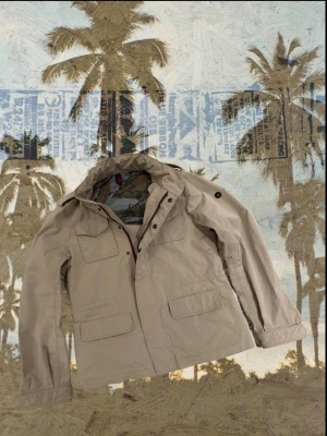 Beige vindjacka med Från Coachrane - Snygg beige vindjacka med camo-mönstrat meshfoder och coola detaljer som röd och marinblå dragkedja. Jackan har flera fickor med lock och praktisk huva. Perfekt för dig som gillar streetstyle och funktionella plagg.