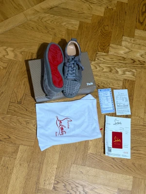 Grå Christian Louboutin sneakers med nitar - Säljer ett par gråa sneakers från Christian Louboutin med coola nitar på tån och klassisk röd sula. Skorna är gjorda i mocka och har snörning samt rund tå. Kommer med originalkartong och dustbag med Louboutin-logga. Och det finns 3 nitar som är borta