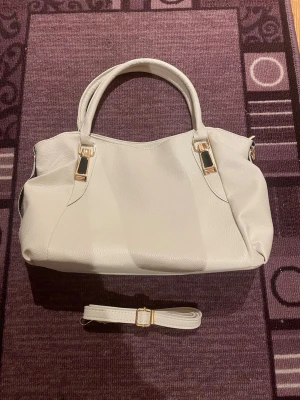 Vit handväska med gulddetaljer - Snygg vit handväska i skinnimitation med guldfärgade metalldetaljer och tillhörande axelrem. Väskan har praktiska handtag, en dragkedja på baksidan och rymligt innerfack. Perfekt för dig som vill ha en stilren och modern look. Perfekt till skolan då den får plats med dator mm. 