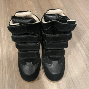 Isabel Marant bekett  - Svarta höga sneakers från Isabel Marant med flera kardborreband och detaljer i mocka och skinn. Skorna har en rund tå, platt sula i gummi och vadderad insida i beige. Perfekt för dig som gillar statement-skor med cool design.