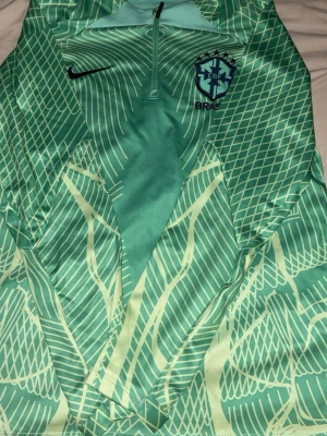 Nike Brasilien träningströja – stil & komfort - Snygg och sportig träningströja från Nike med Brasiliens landslagslogga (CBF). Tröjan har en modern grön design med stilrent grafiskt mönster, broderad Nike-swoosh och halv dragkedja framtill för extra ventilation och komfort.