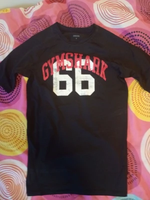 Svart Gymshark t-shirt med tryck - Svart t-shirt från Gymshark i storlek M med stort tryck i rött och vitt på framsidan där det står 'GYMSHARK 66'. T-shirten har korta ärmar och är gjord i mjukt och stretchigt material som passar perfekt till träning. Snygg och sportig stil.