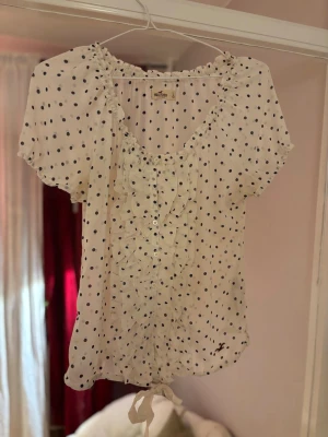 Hollister Blus med prickar - Hollister-blus med prickar i storlek L säljes! Den är vit med mörkblå prickar, har fina volanger framtill och knytdetalj nedtill. Använd men i bra skick. Säljes för endast 80 kr! Passa på!"