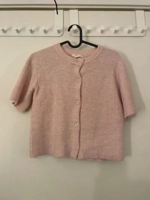 Ljusrosa stickad kofta från Gina Tricot - Säljer en ljusrosa, ribbstickad kofta från Gina Tricot med korta ärmar och rund hals. Koftan har knappar framtill i matchande färg och är gjord i ett mjukt stickat material som känns skönt mot huden. Perfekt för lager-på-lager. 💖💖 