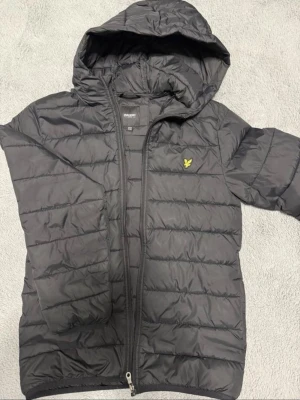 Svart pufferjacka från Lyle & Scott - Snygg svart pufferjacka från Lyle & Scott med gul logga på bröstet. Jackan har huva, dragkedja framtill och quiltad design. Perfekt för kalla dagar och ger en sportig look. Tillverkad i lätt och värmande material.