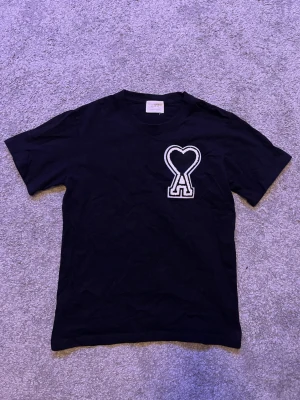 Svart t-shirt från Ami Paris med hjärtlogo - Svart t-shirt från Ami Paris med deras ikoniska hjärtformade A-logo broderad i vitt på bröstet. Klassisk passform med rund hals och korta ärmar. Tillverkad i mjuk bomull som känns skön mot huden.