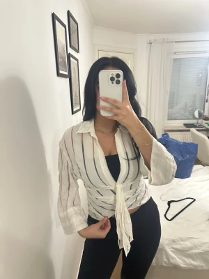 Vit randig skjorta med knyt framtill - Snygg vit skjorta med tunna vertikala ränder i blått och beige. Skjortan har lång ärm, klassisk krage och knyt framtill för en trendig look. Perfekt att styla över ett linne eller topp.