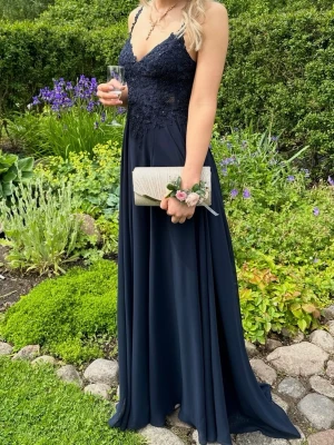 Marinblå balklänning med spets - Elegant marinblå långklänning med spetsdetaljer upptill och tunna axelband. Klänningen har v-ringad urringning och ett flytande, svepande kjolparti. Perfekt för bal. Klänningen har en slits som endast syns när man går. Den har också ett väldigt litet ”släp” som man kan se på sista bilden. Klänningen är använd endast två tillfällen och är kemtvättad efter användning båda gångerna. klänningen är köpt i en lokal balbutik och nypris är ca 5000. Säljer även väskan för 300 kr.