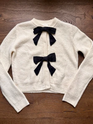 Vit kofta med svarta rosetter - Supersöt vit ribbstickad kofta med två stora svarta rosetter framtill. Koftan har rund halsringning och långa ärmar, perfekt för en trendig och gullig look. Materialet ser mjukt och mysigt ut, och rosetterna ger en extra touch av stil.