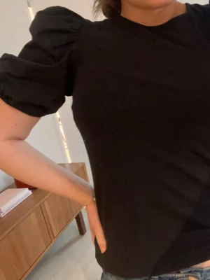 Svart topp med puffärmar - Snygg svart topp med puffiga korta ärmar och rund halsringning. T-shirten har en enkel men trendig design som passar perfekt till jeans eller kjol. Tillverkad i mjukt material för en bekväm känsla.