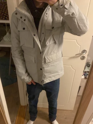 J.lindeberg jacka - Snygg ljusgrå parkas med flera stora fickor framtill och dragkedja med tryckknappar. Jackan har hög krage och huva, perfekt för kyliga dagar. Passar till jeans och har en modern, avslappnad stil.