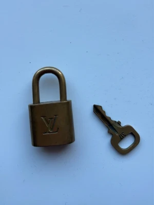 Louis Vuitton hänglås med nyckel - Säljer ett klassiskt mässingsfärgat hänglås från Louis Vuitton med tillhörande nyckel. Låset har LV-loggan ingraverad på framsidan och är perfekt som accessoar till din väska eller ryggsäck. Nyckeln har siffran 317. Snygg detalj för att ge din väska en exklusiv touch.