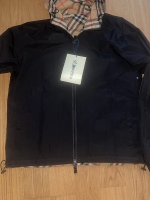 Burberry vindjacka med klassiskt rutmönster - Snygg vindjacka från Burberry med klassiskt beige rutmönster på ena sidan och svart på andra. Jackan är vändbar, har huva och dragkedja framtill. Perfekt för dig som vill ha en stilren och ikonisk look. Materialet är lätt och passar bra till vår och höst.