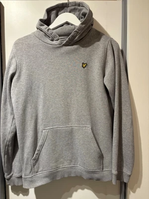 Grå hoodie från Lyle & Scott Junior - Snygg grå hoodie från Lyle & Scott Junior med klassisk gul logga på bröstet. Hoodien har en stor huva, känguruficka framtill och ribbade muddar vid ärmar och nederkant. Tillverkad i mjukt sweatshirtmaterial som är perfekt för chill dagar. Använd max 5gånger