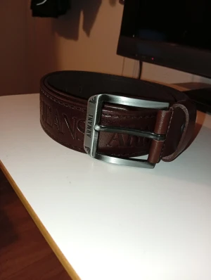 Brunt skärp från Armani Jeans - Snyggt brunt bälte från Armani Jeans i skinn med präglad logga längs hela remmen. Kraftigt spänne i metall med märkets namn ingraverat. Perfekt accessoar för att lyfta din outfit med en exklusiv touch.