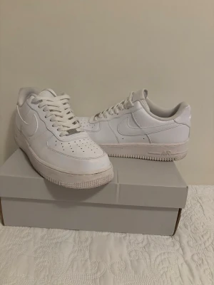 Nike Air Force 1 vita sneakers - Klassiska Nike Air Force 1 sneakers i helvitt läder med perforerad tå, vita skosnören och den ikoniska swoosh-loggan på sidan. Låg modell med bekväm sula och diskret AIR-detalj på mellansulan. Perfekta för en clean och stilren look.