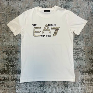 Vit EA7 Emporio Armani t-shirt - Snygg vit t-shirt från EA7 Emporio Armani med tryckt logga och grafisk text på bröstet. Klassisk passform med rund halsringning och korta ärmar. Perfekt för en stilren och trendig look.