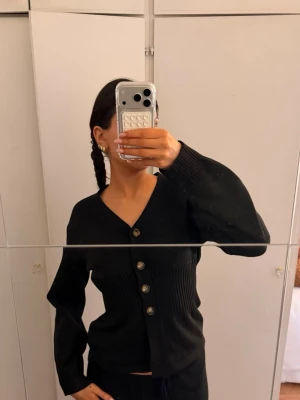 Svart stickad kofta med knappar - Snygg svart stickad kofta med v-ringning och fyra stora dekorativa knappar framtill. Koftan har ribbstickad midja och långa ärmar med lätt ballongeffekt. Perfekt att styla till jeans eller kjol. Använd 2 ggr.