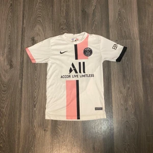 PSG matchtröja vit rosa Nike - Säljer en snygg PSG matchtröja från Nike i vitt med rosa och svart rand på framsidan. Tröjan har korta ärmar, klubbmärke på bröstet och sponsorlogga. Perfekt för dig som älskar fotboll och vill ha en unik look på planen.
