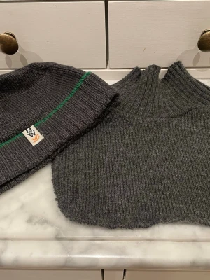 Mörkgrå ribbad mössa från Lindex - Snygg mörkgrå ribbstickad mössa från Lindex med en grön rand och liten etikett framtill. Materialmix med akryl, polyester och ull för extra värme och komfort. Storlek 56/58