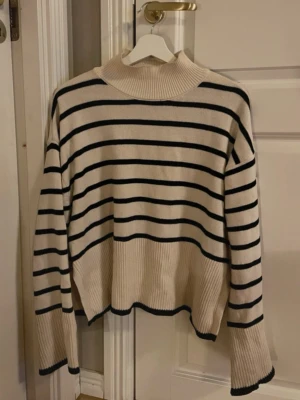 Randig stickad tröja Gina Tricot - Säljer en krämvit stickad tröja från Gina Tricot med svarta horisontella ränder. Tröjan har hög krage, långa ärmar och ribbade muddar. Den har en avslappnad passform. Perfekt för dig som gillar klassisk stil.