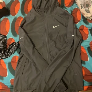 Mörkgrå Nike vindjacka med huva - Snygg mörkgrå vindjacka från Nike med hel dragkedja, huva och två fickor med dragkedja. Jackan har en minimalistisk design med Nike-logga på bröstet och diskreta reflexdetaljer på ärmarna. Perfekt för blåsiga dagar och sportiga outfits.
