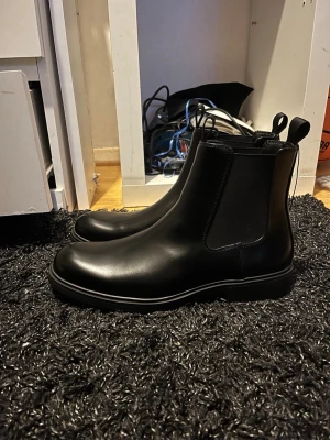 Svarta Chelsea boots i skinn - Säljer ett par stilrena svarta Chelsea boots i slätt skinn med elastiska paneler på sidorna och dragflikar både fram och bak. Klassisk siluett med rundad tå och låg sula, perfekta för en clean och modern look. Endast testade säljer för att de var för stora 