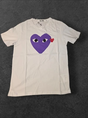Vit Comme des Garçons PLAY t-shirt - Vit t-shirt från Comme des Garçons PLAY med ett stort lila hjärta med ögon och ett litet rött hjärta på bröstet. Klassisk passform och rund hals. Perfekt statement-plagg för dig som gillar streetwear och grafiska prints.